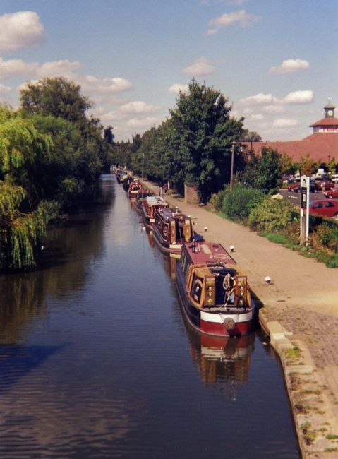 A serene waterfront scene along the Grand Union Canal featuring a paved promenade on the right with a low brick edging, lined with lush green trees and shrubs. In the foreground, reflective canal wate